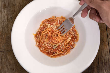 A hand holding a fork inside a white plate of ready-to-eat pasta with bolognese sauce and cheese on...