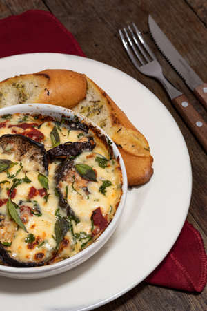 Ready-to-eat lasagna with eggplant, mozzarella, parmesan, basil, parsley and garlic bread in a white plate on a wooden background and next to some cutlery