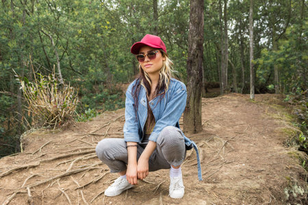 A young white woman wearing a denim jacket crouching looking at the camera in a forest on a sunny...
