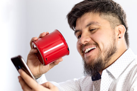 A young white man with an earring smiling while checking his cell phone and drinking coffee from a...