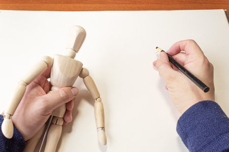 Point of view of an artist holding a drawing mannequin with his left hand while drawing it with his...