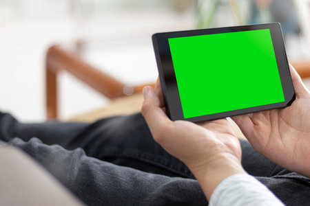 Man holding a black tablet with chroma green screen while lying on a sofa at home. A person watching...