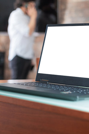 A laptop with a blank screen on a work desk with an unrecognizable person talking on a cell phone out of focus in the background. Home office space.の写真素材