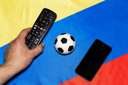 Hand holding a remote control next to a soccer ball and smartphone on a background with the colors...
