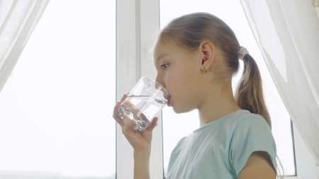 Girl teenager drinking clear water from glass on home kitchen on window background. Thirsty girl...
