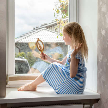 School girl combs her hair sitting on the windowsill, rainy weather outsideの写真素材