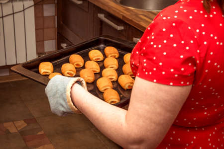 A woman carries cooked buns on a baking sheet. The woman in the kitchen made bread rolls.