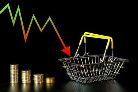 Shrinking stacks of coins and an empty grocery basket on a black background, an arrow showing the economy fallingの写真素材