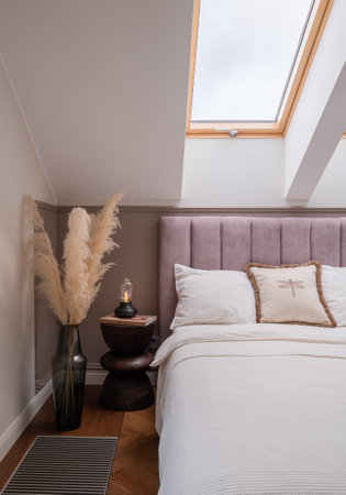 A bedroom with a bed, nightstand, vase of pampas grass, and a skylight fixtureの写真素材