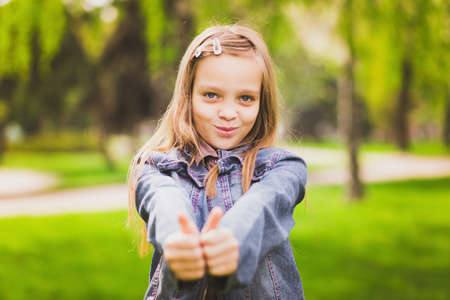Young girl puts her big thumbs up. Portrait of cheerful kid. Smiling girl having fun outside. Happy successful person.の写真素材