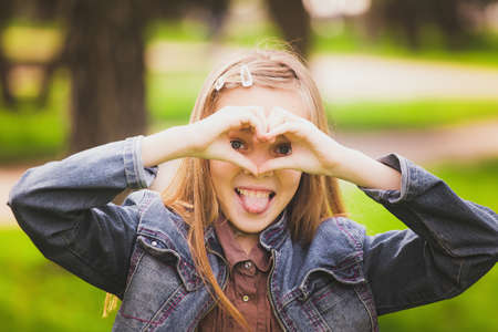 Portrait of young girl with tongue out  having fun outside. Funny kid looking cheerfully at camera through heart made by hands. Horizontal color photo.の写真素材