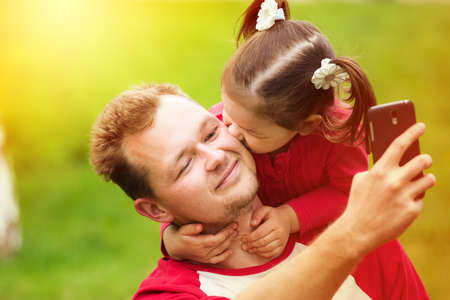 Portrait of a little girl kissing her father on cheek while taking selfie with phone. Close up face...