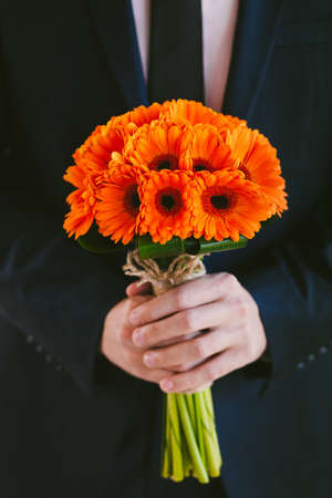 Groom in blue suit holding wedding bouquet of flowers in hands waiting for his bride. Boutonniere and bridal bouquet of orange gerbera daisies.の写真素材