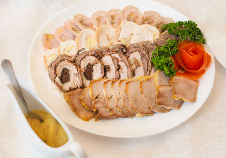 Close up of white plate full of meat appetizers decorated with red rose made of tomato and fresh leaves of parsley. Assortment of sliced meat, chicken and pork rolls. Saucepan with sauce.の写真素材