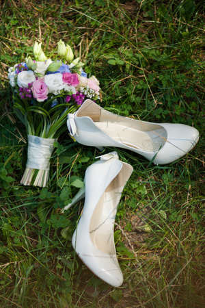 Pair of white ladies shoes on grass and beautiful bouquet of flowers. Wedding accessories.の写真素材