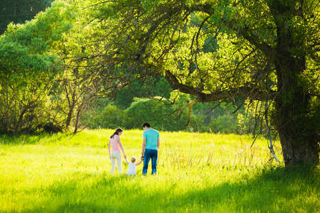 Family of mum, dad and little daughter walking together in beautiful meadow. Silhouettes of young parents holding hands of their baby girl. First steps of child in long grass.の写真素材