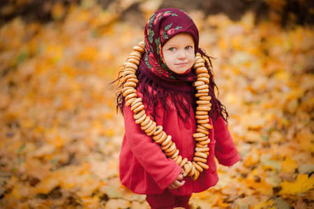 Child girl in Ukrainian folk scarf on head with floral print and with bunch of bagels on background of fallen leavesの写真素材