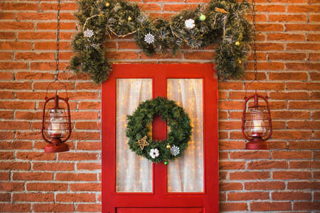Christmas decor. Festively decorated interior in red, brown colors. Horizontal colour image of photo booth with brick wall, painted red door, hanging lanterns, christmas garland and pine border.の写真素材