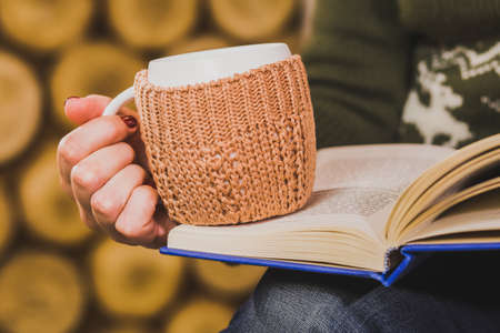 Cup of hot tea or coffee or cocoa in knitted vintage cup holder with christmas decorations in hands of woman with beautiful festive red manicure. Woman sitting in rustic interior and reading bookの写真素材