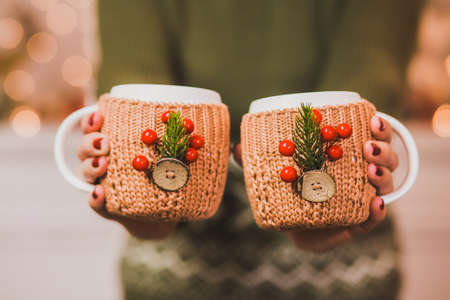 Cute winter Christmas mugs. Cups of hot tea or coffee or cocoa in knitted vintage cup holders with xmas decorations in hands of woman with festive red manicure. Woman holding cups together closely.の写真素材