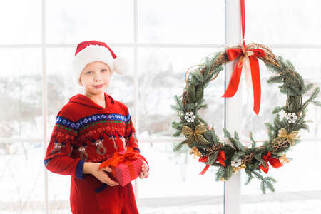 Portrait of cute little child in holiday christmas interior. Happy smiling kid posing for photo standing near big window with snowy city landscape seen through it. Horizontal color photography.の写真素材