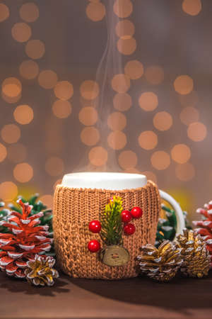 Closeup of white steaming winter cup in brown knitted sweater with christmas decorations on wooden...
