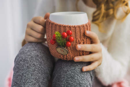 Close up of hands of child holding white mug in winter knitted cover festively decorated with Christmas details. Cute little girl sitting on windowsill, drinking hot tea. Horizontal color photoの写真素材