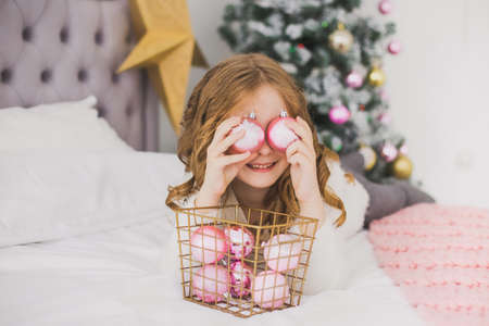 Portrait of cute happy little girl on Christmas morning in home interior. Child paying with holiday decorations, making eye of pink balls while laying on bed. Horizontal color photoの写真素材