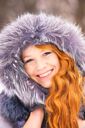 Portrait of beautiful young girl dressed in fur coat at winter trees background. Beauty woman having fun outside on snowy day. Close up of female smiling face with gorgeous long ginger hair.の写真素材