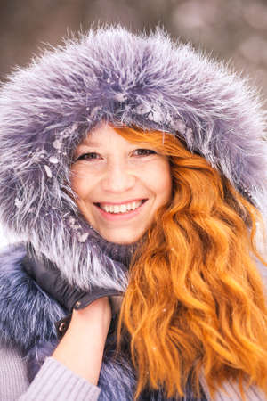 Portrait of smiling ginger winter girl looking cheerfully at camera. Close up of beauty face of young woman in winter fur clothing outside at snowy cold winter day backgroundの写真素材