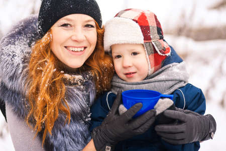 Close up portrait of baby sitting with young beautiful mother outside at snowy trees winter background and drinking hot tee. Happy family enjoying beautiful frosty days on picnic Horizontal photo.の写真素材
