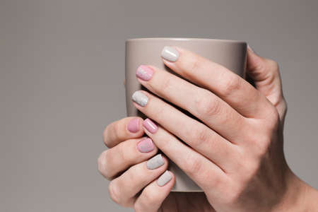 Closeup view of white hands of healthy woman with beautiful modern grey and pink manicure design. Woman holding brown cup. Horizontal color photography.の写真素材