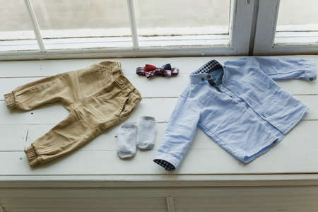 Set of baby clothes. Blue shirt, brown pants, white socks and red bowtie laying on white windowsill....