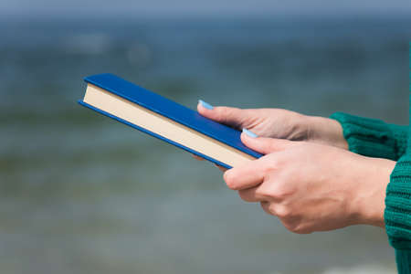 Closeup of white female hands  holding thick paper book in hands isolated at blurry blue sea backgroundの写真素材