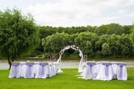 Wedding decor in white and purple colors outdoors on green lawn near river. Scenic place for romantic marriage celebration. Horizontal color photography.の写真素材
