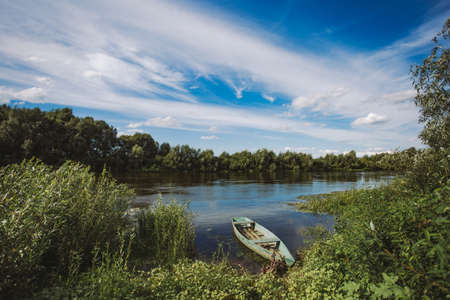 One small lonely empty wooden old boat standing at river bank in beautiful summer countryside landscape. Green trees in background. Horizontal color photography.の写真素材