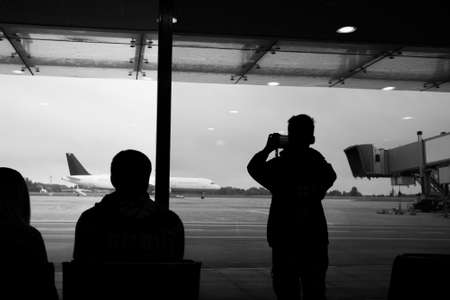 Black and white picture of family waiting their flight at international airport. Child takes photo of huge plane through window of terminal.の写真素材