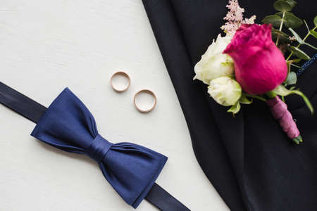 Close up top view of elegant modern formal male accessories. Blue bowtie, two golden wedding rings, flower boutonniere pinned to black jacket of suit. Set for wedding isolated on white background.の写真素材
