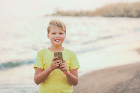 Portrait of cute happy smiling white kid using modern smart phone standing at sunset sandy beach of hotel resort in summer. Happy holidays and vacations concept. Horizontal color photography.の写真素材