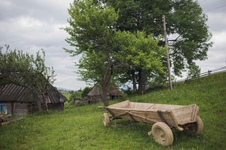 Scenic countryside landscape. Old brown house, barn and wooden cart situated on green grassy hill in...