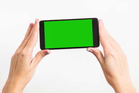 Close up mockup image of woman's two hands holding black mobile phone with blank green screen isolated on white background. Horizontal color photography.の写真素材