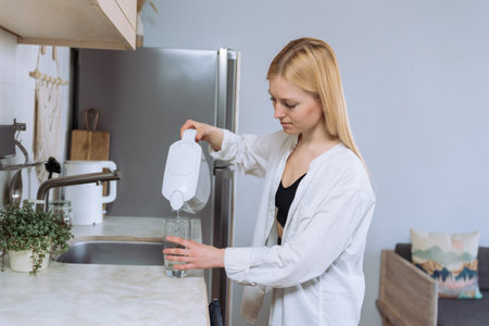 A beautiful young woman in a white shirt pours clean water from a filter in the kitchenの写真素材