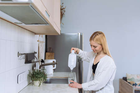 A young woman pours clean water into a glass in a bright and clean kitchen
