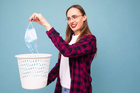 A young woman throws a used protective mask in the trash on a blue background