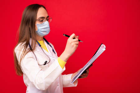 A young woman in a white medical coat and a protective mask writes down a statement in a notebookの写真素材