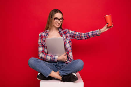 A pretty young woman in stylish clothes is sitting with a paper cup and a laptop on a red backgroundの写真素材