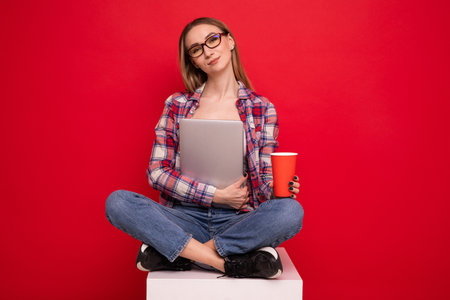 A pretty young woman in stylish clothes is sitting with a paper cup and a laptop on a red backgroundの写真素材