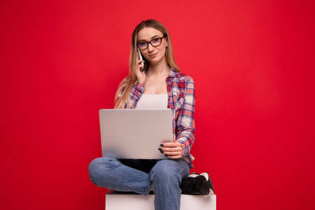 Cute young woman in stylish clothes talking on the phone and working with a laptop on a red backgroundの写真素材