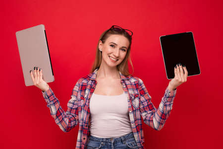 A pretty young woman in stylish clothes holds a tablet and a laptop on a red backgroundの写真素材