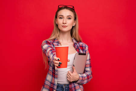 A pretty young woman in stylish clothes holds a tablet and a paper cup for coffee on a red backgroundの写真素材